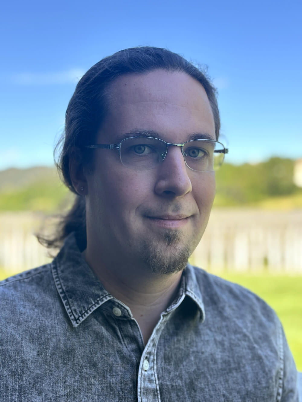 headshot of brandon hubler. 30's white male with glasses, goatee and ponytail, wearing black button up shirt. background is a blurry fenceline and mountain